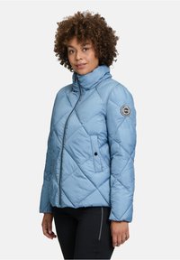 Błękitna pikowana kurtka z wysokim kołnierzem, pełnym zapięciem na zamek i bocznymi kieszeniami. Posiada naszywkę z logo na lewym rękawie oraz gładką teksturę.