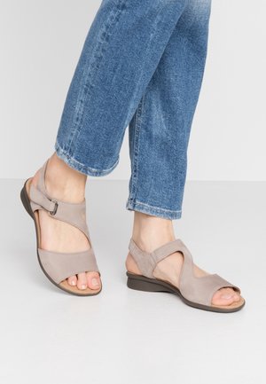 Fødder iført beige åbne sandaletter med justerbare stropper sammen med blå denimjeans på en hvid gulv og neutral baggrund.