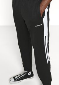 Black Adidas sweatpants with white side stripes, featuring an elastic waistband and pockets, worn with black and white sneakers.