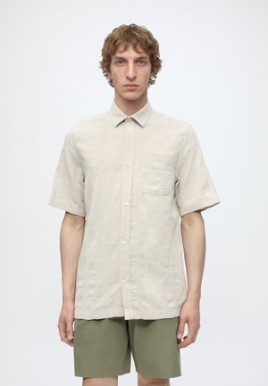Jeune homme aux cheveux bouclés portant une chemise beige à manches courtes boutonnée et un short vert olive, debout sur un fond blanc uni.