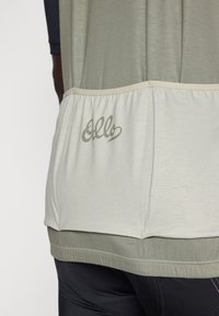Camisa atlética gris con una cintura fruncida en gris claro, que presenta el logo "Ello" en una impresión sutil. Tejido suave y texturizado en toda la prenda.