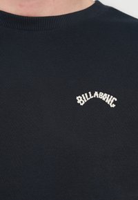 Tengerészkék pulóver bordázott nyakkivágással és egyszerű, fehér "Billabong" hímzett logóval a mellkason. Puha tapintású, normál szabású.
