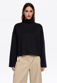 Zwarte cropped top met een hoge kraag, lange mouwen en een gladde textuur. Heeft een relaxte pasvorm en een minimalistisch ontwerp. Gepaard met beige broek.