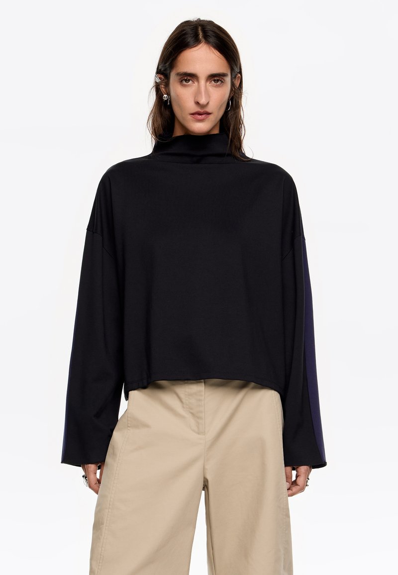 Zwarte cropped top met een hoge kraag, lange mouwen en een gladde textuur. Heeft een relaxte pasvorm en een minimalistisch ontwerp. Gepaard met beige broek.