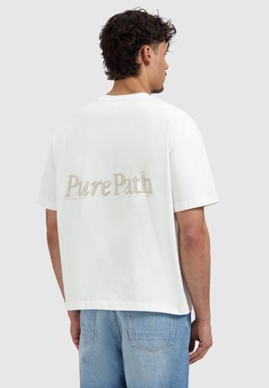 Mann mit lockigem dunklem Haar, der ein weißes oversized T-Shirt mit dem Schriftzug „Pure Path“ auf dem Rücken und hellblaue Jeans trägt, steht mit dem Rücken zur Kamera.