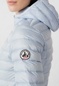 Lichtblauwe donsjas met capuchon, voorzien van horizontale stiksels, een ronde patch op de mouw en een gladde, glanzende textuur.