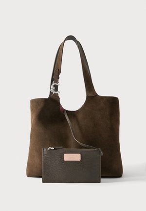 Sac fourre-tout en daim marron avec sangle réglable et pochette zippée en cuir foncé attachée, avec une étiquette de marque en métal à l’avant.