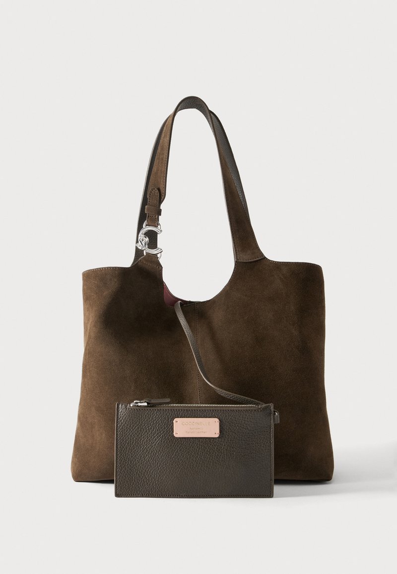 Sac fourre-tout en daim marron avec sangle réglable et pochette zippée en cuir foncé attachée, avec une étiquette de marque en métal à l’avant.