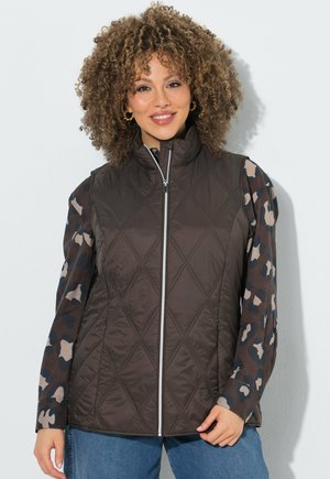 Donna sorridente con capelli ricci che indossa orecchini a cerchio dorati, un gilet trapuntato marrone scuro con cerniera, una maglia mimetica a maniche lunghe e jeans blu davanti a un muro bianco.