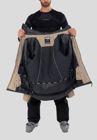 Béžová a tmavě modrá lyžařská bunda s kapucí, která má několik kapes a hladké vnitřní podšívku, držena otevřená, aby ukázala detaily.