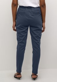 Personne portant un pantalon tapered bleu marine avec des poches arrière et des sandales à petit talon noires, se tenant devant un fond clair uni.