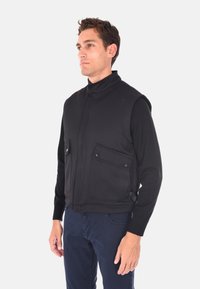 Gilet nero con colletto a costine, due tasche a patta e tessuto testurizzato. Indossato sopra una camicia a maniche lunghe, presenta un design aderente.