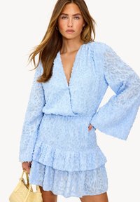 Robe en dentelle bleu clair avec de longues manches en cloche, décolleté en V profond, ourlet à volants et motif floral texturé. Associée à un sac à main jaune.