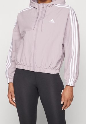 Lila Adidas Kapuzenjacke zum Zuziehen mit weißen Streifen an den Ärmeln, getragen zusammen mit schwarzen Leggings an einer stehenden Person.