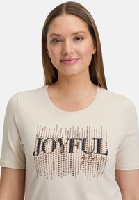 T-shirt beige clair à manches courtes, ornée de clous métalliques disposés en motif vertical, épelant "JOYFUL" en texte noir.
