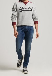 Persona caminando con un sudadera gris de SuperDry con puños color burdeos, jeans azules y zapatillas altas negras con cordones blancos.