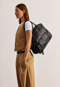 Zaino in pelle nera con molteplici scomparti con cerniera, forma arrotondata e manici robusti. Abbinato a un gilet e pantaloni beige.