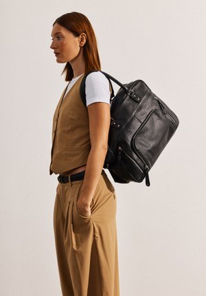 Zaino in pelle nera con molteplici scomparti con cerniera, forma arrotondata e manici robusti. Abbinato a un gilet e pantaloni beige.