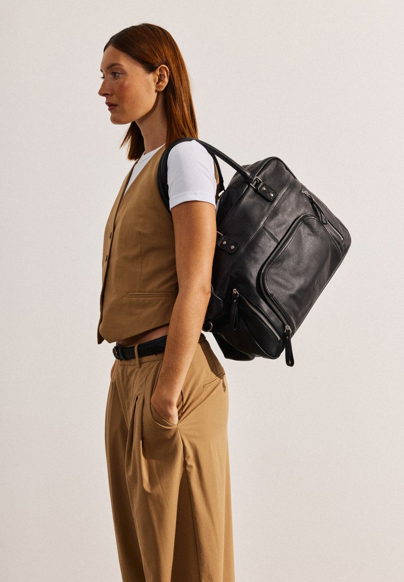 Zaino in pelle nera con molteplici scomparti con cerniera, forma arrotondata e manici robusti. Abbinato a un gilet e pantaloni beige.