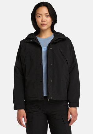 Zwart, hoodie-jacket met drukknoopjes en zakken, gedragen over een lichtblauw overhemd. De stof lijkt duurzaam en weersbestendig.