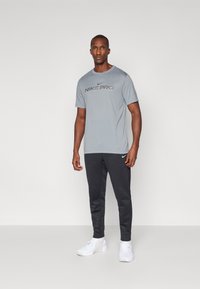 Nike Pro grå t-skjorte med svart logo, kombinert med svarte joggebukser og hvite sko. Glatt stoff, crew neck-design og tettsittende silhuett.