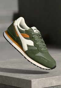 Green athletic shoe with white and orange accents, featuring a textured upper, laces, and a white sole with a black rubber base.