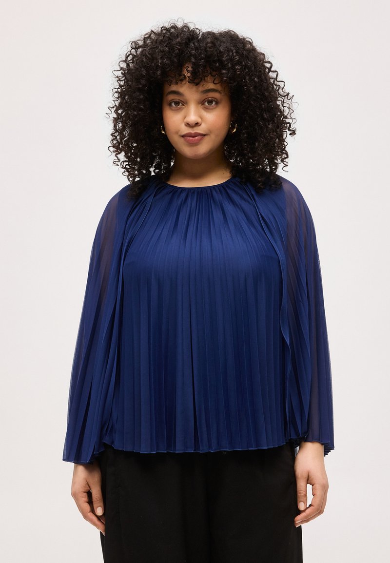 Γυναίκα με σγουρά μαλλιά που φοράει μπλε μπλούζα πλισέ με μακριά μανίκια και μαύρο παντελόνι, στέκεται μπροστά σε απλό φωτεινό φόντο.