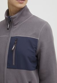 Grijze fleecejack met een marineblauwe nylon borstpaneel, hoge kraag en een ritszak. Voorzien van branding op de ritsen en textuurstof.