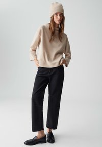 Maglione a maglia beige con maniche corte, abbinato a pantaloni neri a gamba larga e mocassini neri. Il modello indossa un berretto beige coordinato.