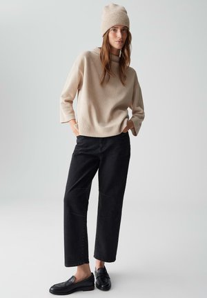 Maglione a maglia beige con maniche corte, abbinato a pantaloni neri a gamba larga e mocassini neri. Il modello indossa un berretto beige coordinato.