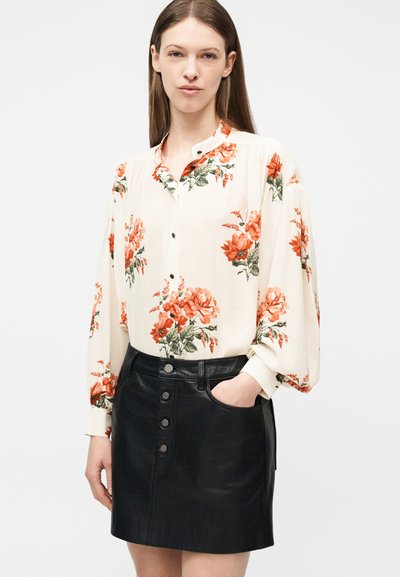 Γυναίκα που φοράει κρεμ μπλούζα με πορτοκαλί λουλούδια και μαύρη δερμάτινη φούστα με κουμπιά μπροστά, με το ένα χέρι στην τσέπη, ουδέτερο φόντο.