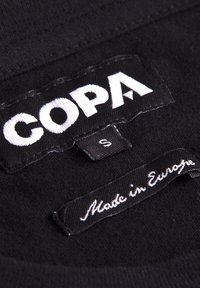 Zwarte stoffen label met "COPA" in vetgedrukte witte letters en maat "S". Een ander label leest "Made in Europe" in cursieve witte tekst.
