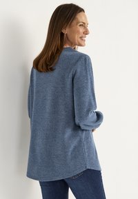 Lächelnde Frau mit glattem braunem Haar, die einen blauen gerippten Pullover und dunkelblaue Jeans trägt und seitlich vor einer schlichten weißen Wand posiert.