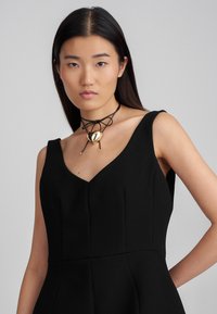 Μαύρο εφαρμοστό φόρεμα με V-ντεκολτέ, που διαθέτει δομημένα πάνελ και ανοίγματα για τα χέρια. Φορεμένο με μαύρο κορδόνι χεριού διακοσμημένο με χρυσό κρεμαστό κ heart pendant.