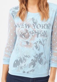 Top de malla azul claro con puntos en relieve, estampado floral y texto impreso "NUEVA YORK FASHION". Puños y dobladillo ajustados.