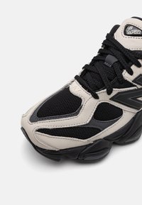 Gros plan d'une basket New Balance 9060 noire et beige montrant des matériaux en mesh et cuir avec des lacets noirs et une semelle en caoutchouc.