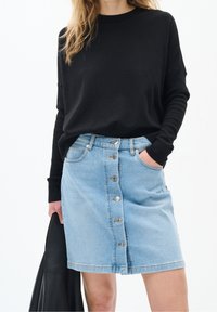 Kvinna som bär en svart långärmad topp och en ljusblå knapp-front denimkjol, hållande en svart läderjacka i ena handen.