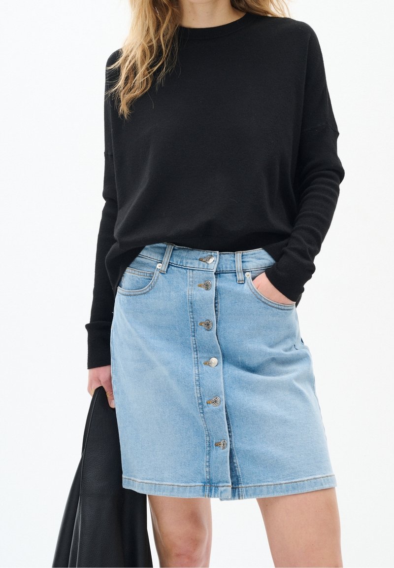 Kvinna som bär en svart långärmad topp och en ljusblå knapp-front denimkjol, hållande en svart läderjacka i ena handen.