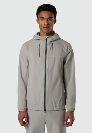 Mann trägt eine hellgraue Kapuzenjacke mit Frontreißverschluss und passende Hose, steht vor hellem, einfarbigem Hintergrund.