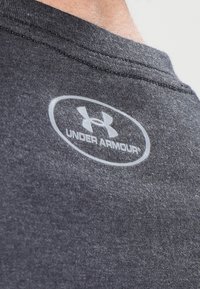 Grå bomullströja med en slät textur och en synlig vit oval logotyp med texten "UNDER ARMOUR" placerad vid halsringningen.