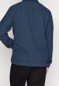 Giacca a quadri blu caratterizzata da un tessuto testurizzato, polsini con bottone e collo a punta, con una parte posteriore visibile e pantaloni scuri.