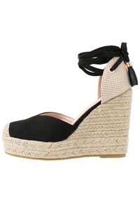 Espadrille compensée en daim noir avec semelle tissée en jute. Dotée d'un dessus en jute beige et d'un lacet noir à la cheville, elle améliore le soutien et le design.
