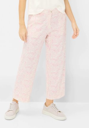 Femme portant un pantalon large à motifs de feuilles rose clair et blanc, un haut blanc et des baskets beiges, debout devant un fond blanc.