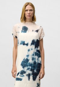 Vrouw met schouderlang rood haar, gekleed in een wit jurkje met korte mouwen en donkerblauwe abstracte patronen, staande tegen een effen lichte achtergrond.
