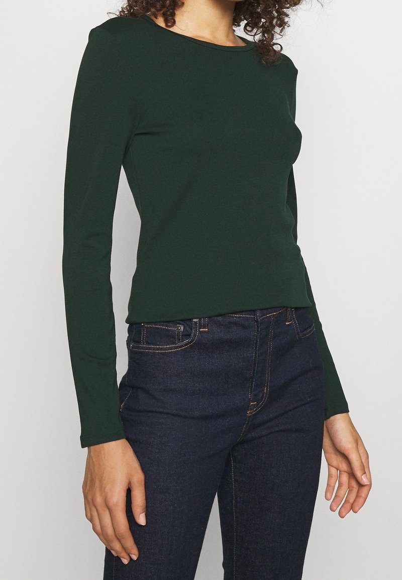 Grön långärmad åtsittande topp i bomullsblandning, med en klassisk rund halsringning och slät yta, tillsammans med mörkblå jeans.