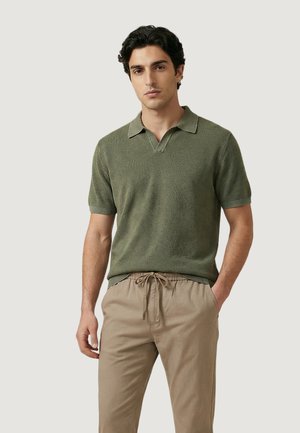 Uomo con capelli scuri che indossa una polo verde oliva a maniche corte e pantaloni beige con coulisse, in piedi contro uno sfondo chiaro e semplice.