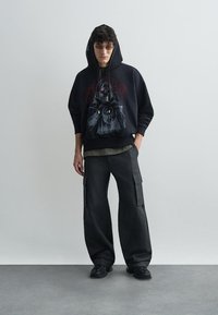 Hanorac negru cu design grafic, asociat cu pantaloni cargo largi și închiși la culoare. Ținuta este completată cu adidași negri și ochelari de soare.