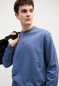 Modrá mikina s textúrovaným povrchom, rebrovaným výstrihom a malým čiernym logom "BOSS" na hrudníku. Model drží čiernu tašku.