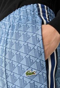 Lyseblå mønstrede joggebukser med et teksturert geometrisk design, elastisk midje og marineblå stripete detaljer. Har en grønn brodert logo.