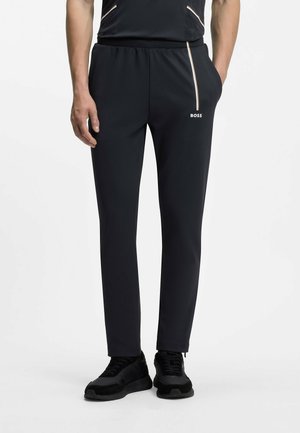 Mann trägt schwarze Slim-Fit-Hose mit weißem Seitenstreifen und "BOSS"-Logo, kombiniert mit schwarzen Sneakers, steht mit den Händen in den Taschen.